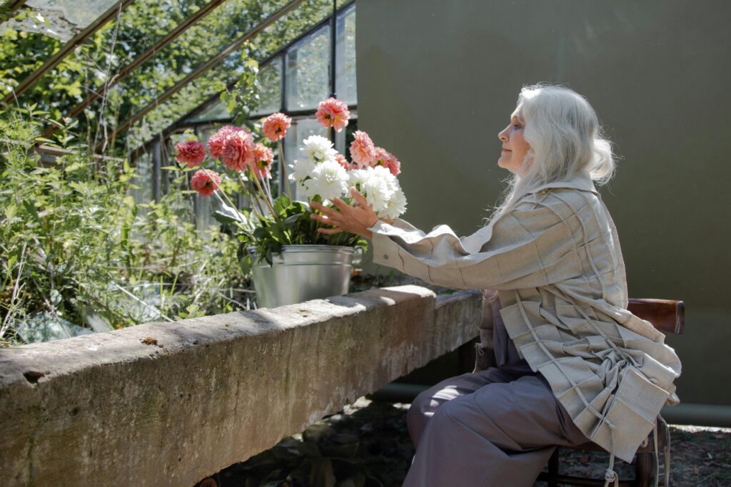 A senior woman with gray hair arranges flowers in a sunny greenhouse setting.